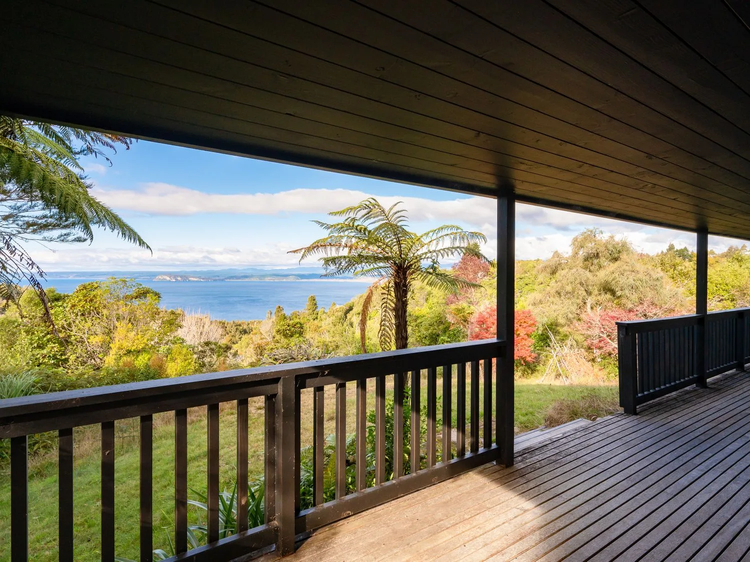 A covered wooden deck overlooking trees and a lake with distant hills at Lake View Lodge - Taupo Holiday Home in Pukawa Bay