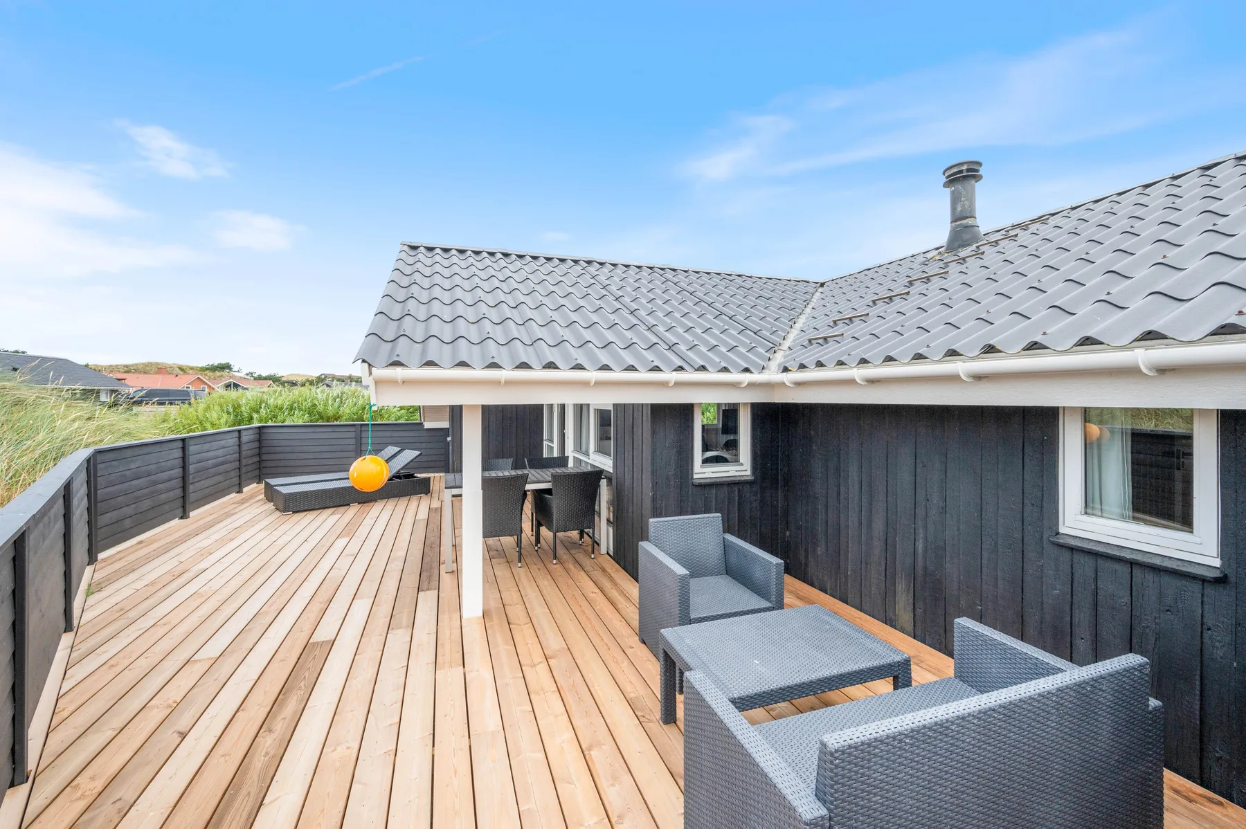 Una terraza de madera junto a una casa de revestimiento oscuro tiene asientos exteriores y un borde cercado.
