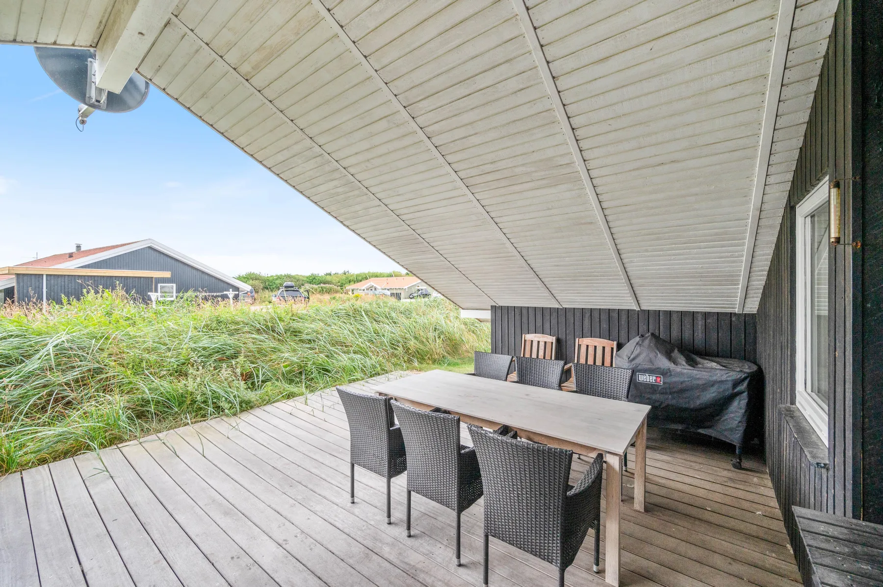 Una terraza exterior cubierta tiene una mesa de comedor y asientos junto a hierba alta.