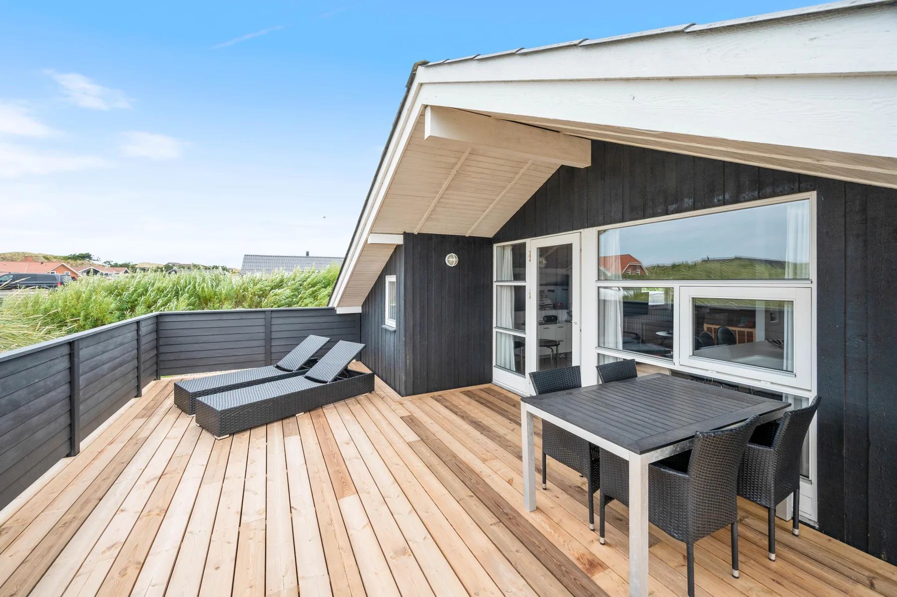 Una terraza de madera junto a una casa de revestimiento oscuro tiene asientos y una mesa de comedor.