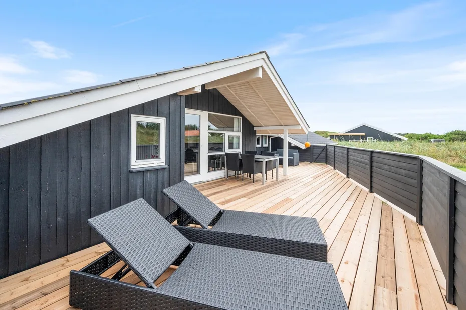 Una terraza de madera junto a una casa de fachada oscura tiene asientos y un comedor.
