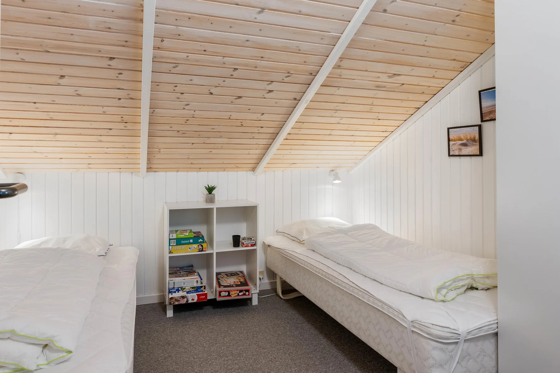 Un dormitorio pequeño con dos camas y una estantería bajo un techo inclinado de madera.