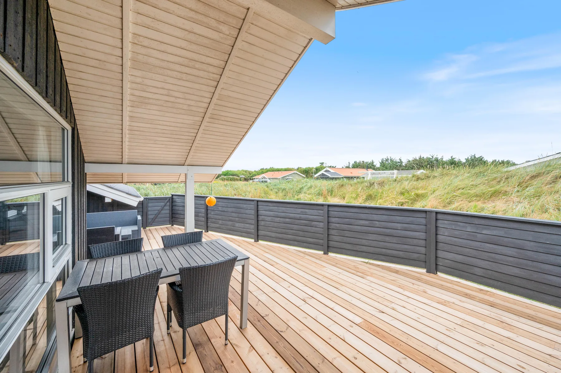 Una terraza de madera cubierta tiene asientos exteriores y una barandilla oscura frente a dunas cubiertas de hierba.