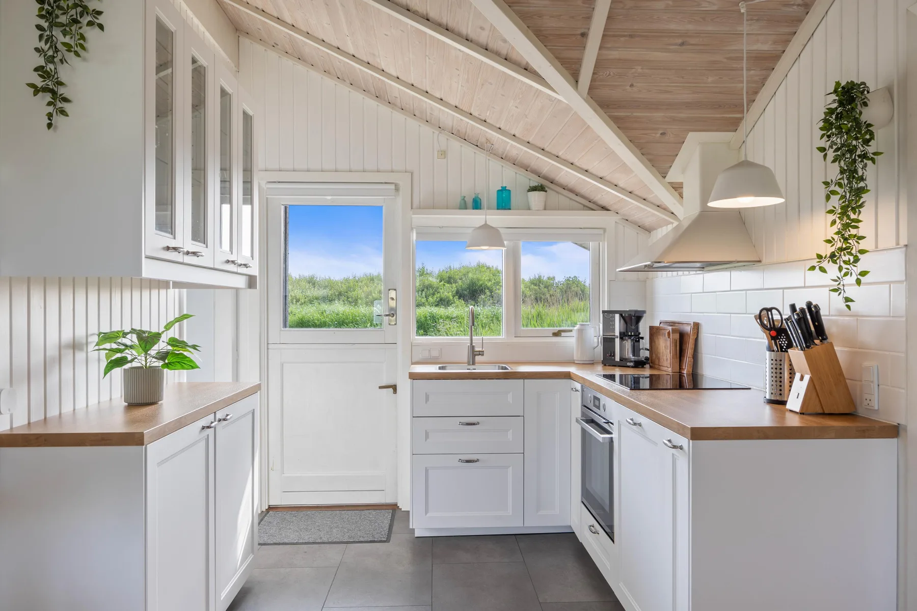 Una cocina luminosa con armarios blancos, encimeras de madera y ventanas con vistas a la vegetación.