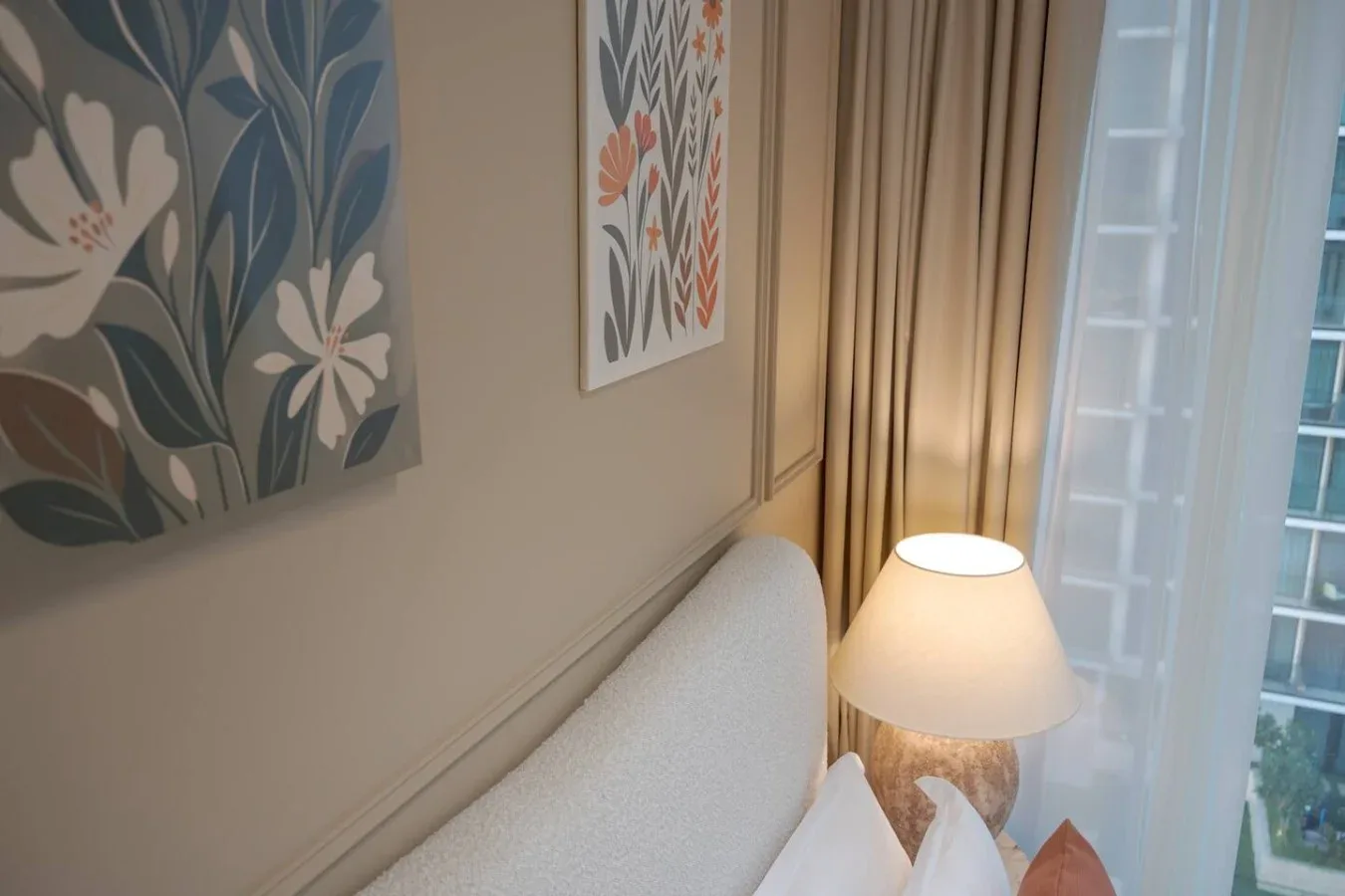Una esquina de dormitorio con cama, lámpara de noche, cortinas y arte floral en la pared.