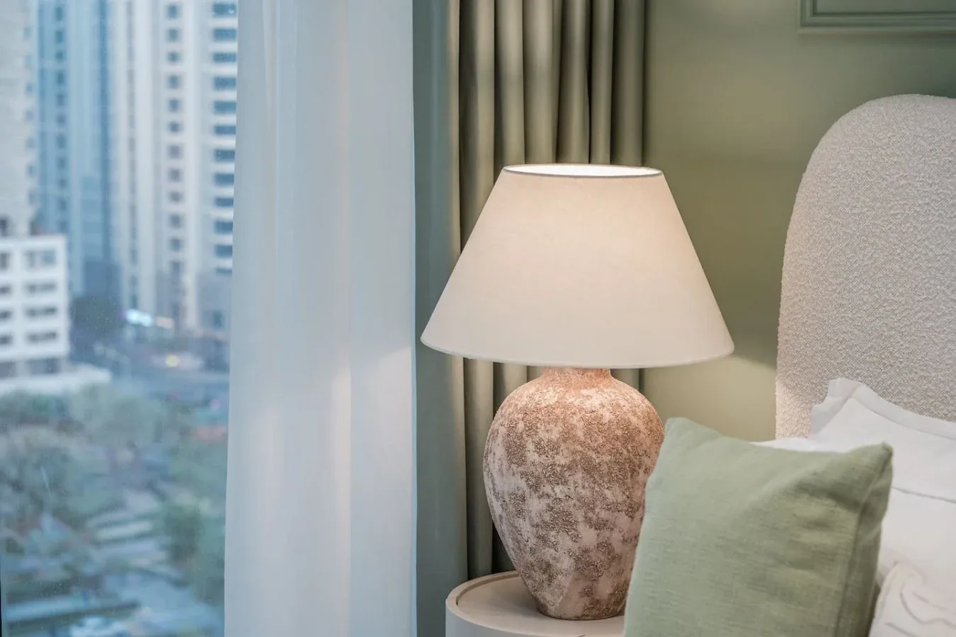 Un rincón de dormitorio con una lámpara de noche y una ventana con vistas a edificios cercanos.