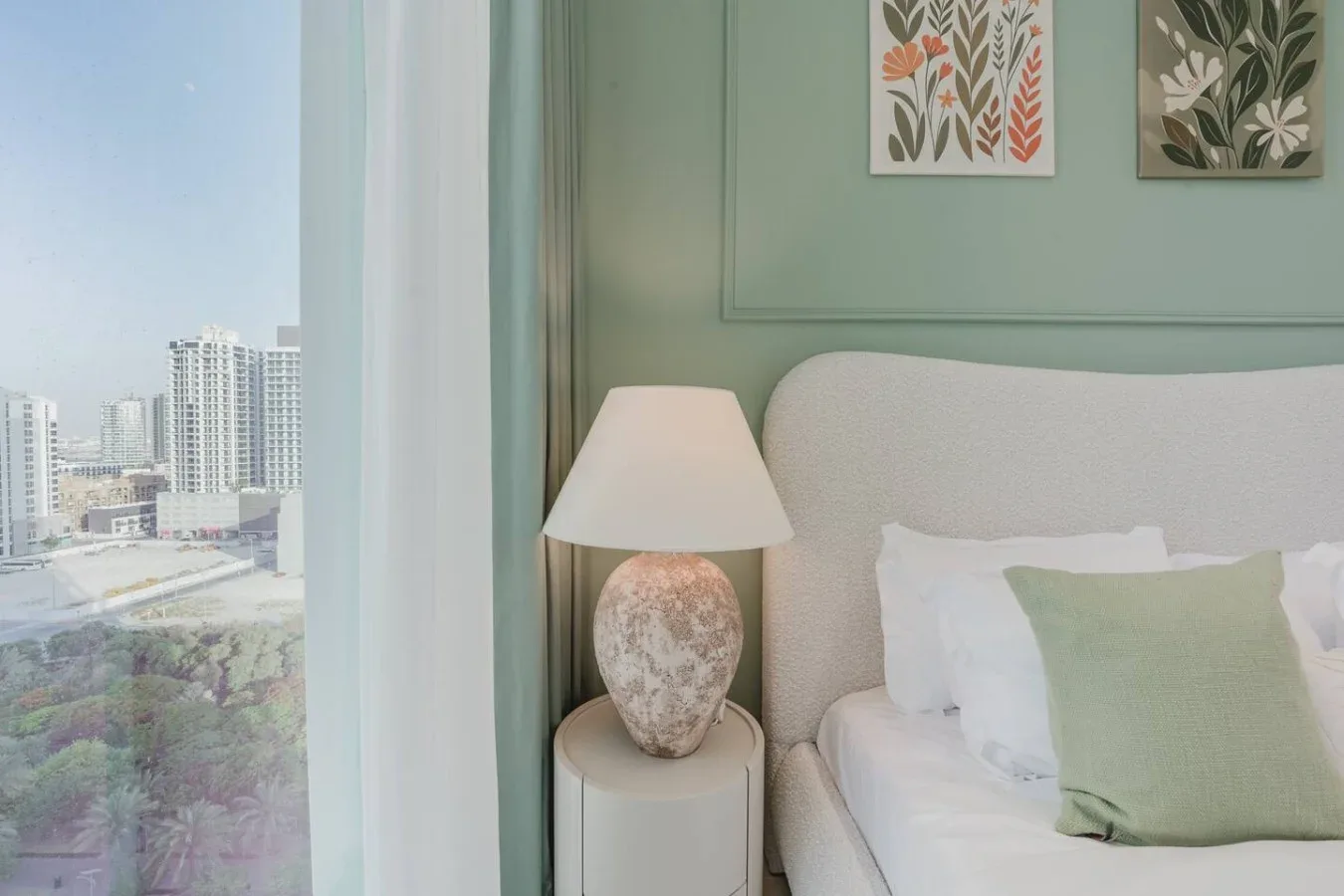 Un dormitorio con cama, lámpara de noche y una gran ventana con vista a edificios cercanos.