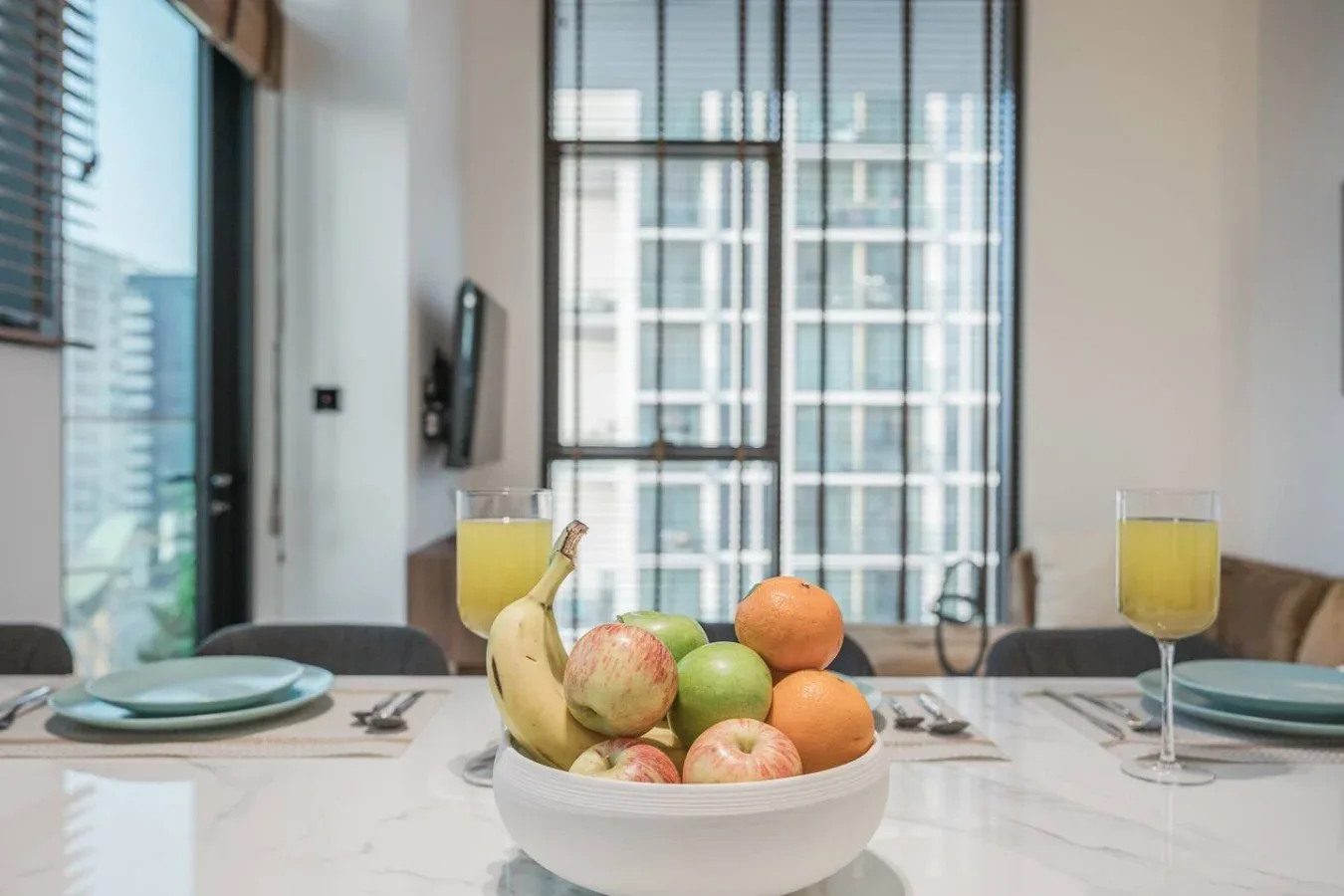 Una mesa de comedor con un frutero y cubiertos está frente a grandes ventanas.