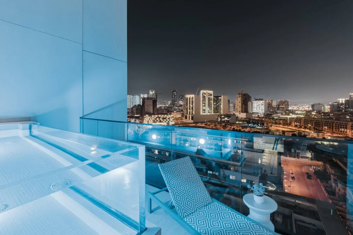 Una terraza en la azotea por la noche ofrece vistas del horizonte urbano, con una silla junto a la barandilla de vidrio.