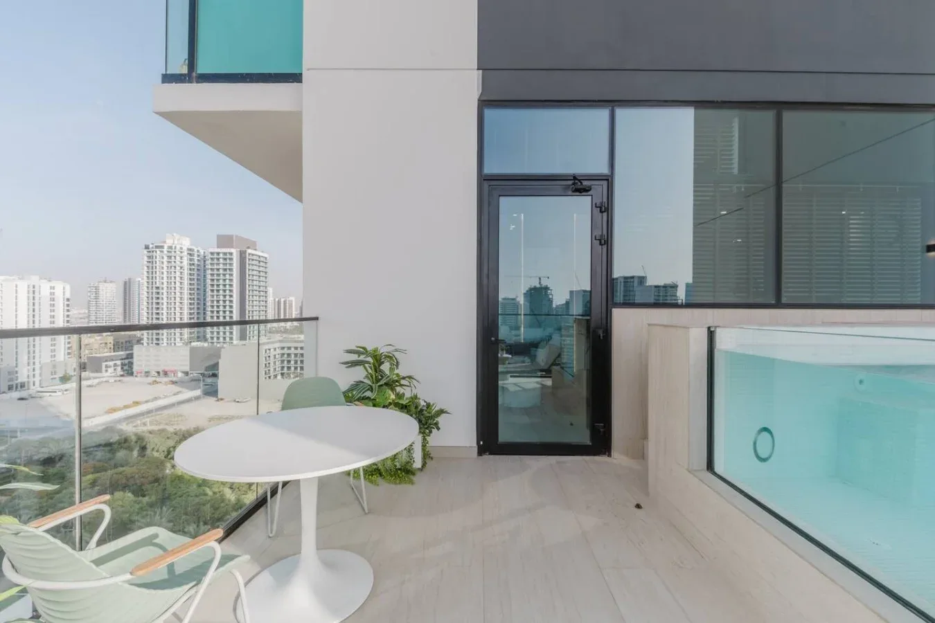 Un balcón moderno con barandilla de vidrio, asientos al aire libre y vista a edificios cercanos.