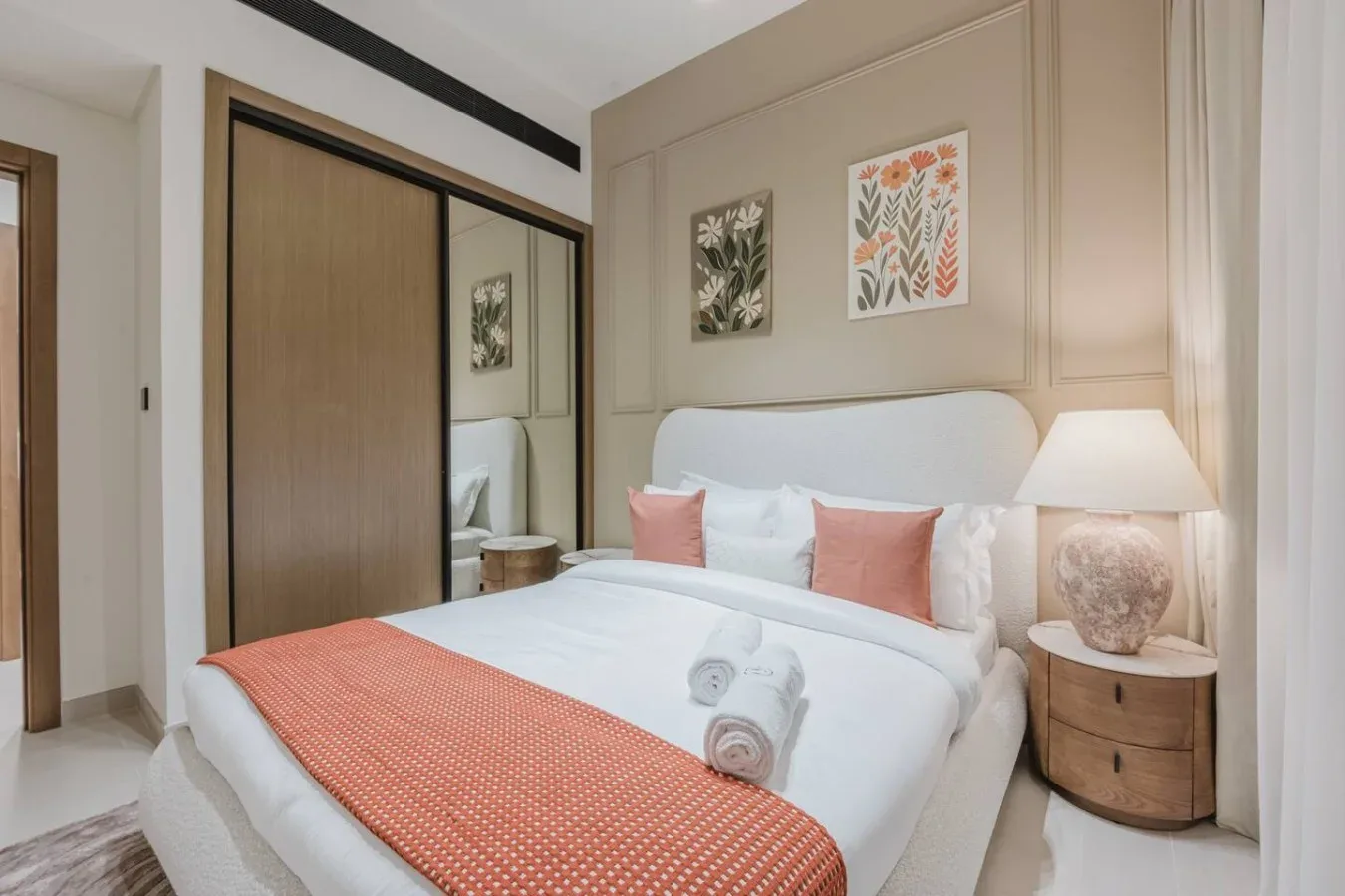 Un dormitorio con una cama tendida, cojines coral y láminas botánicas enmarcadas.