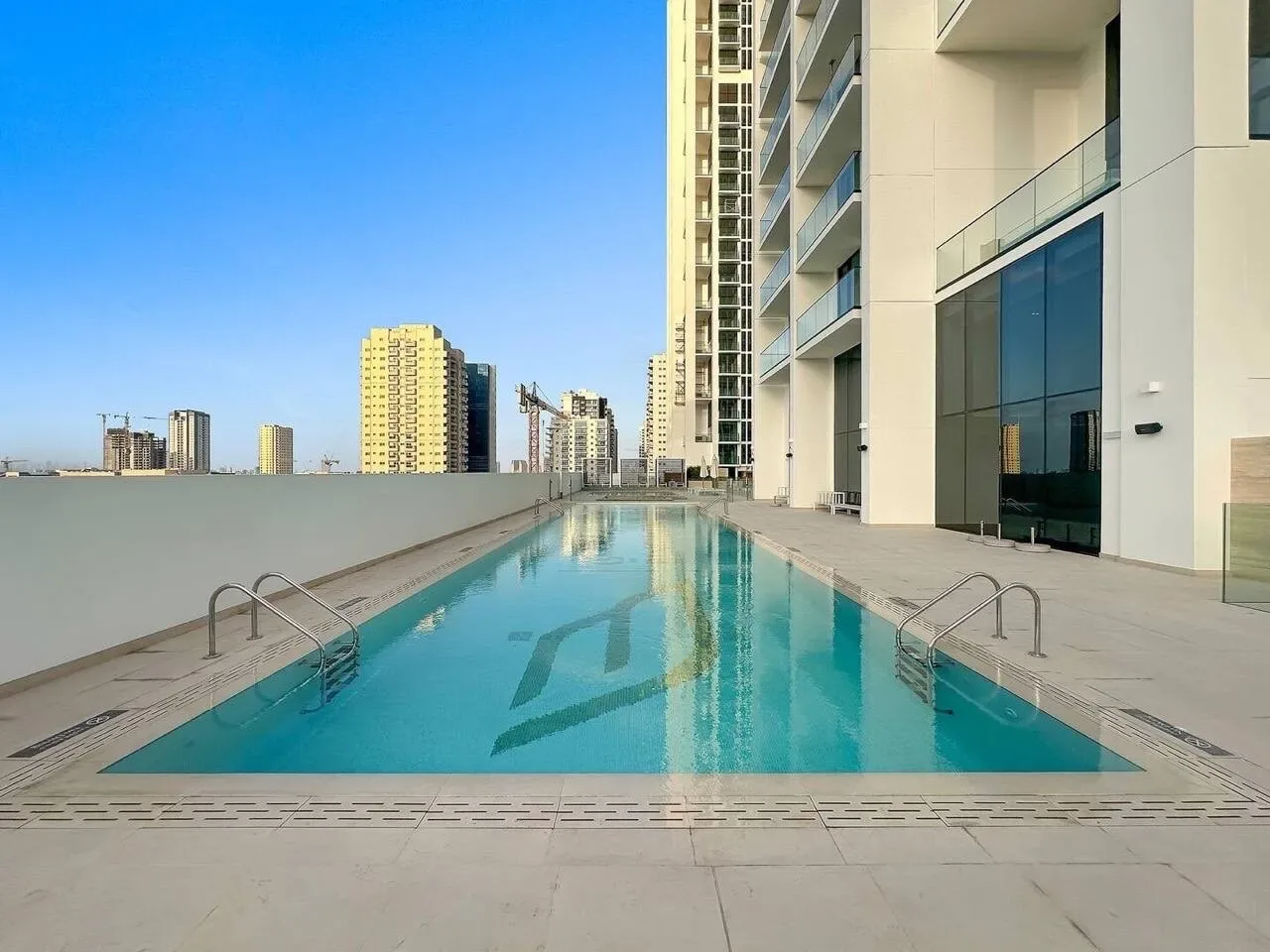 A rooftop swimming pool sits beside a modern high-rise, with city buildings visible beyond.