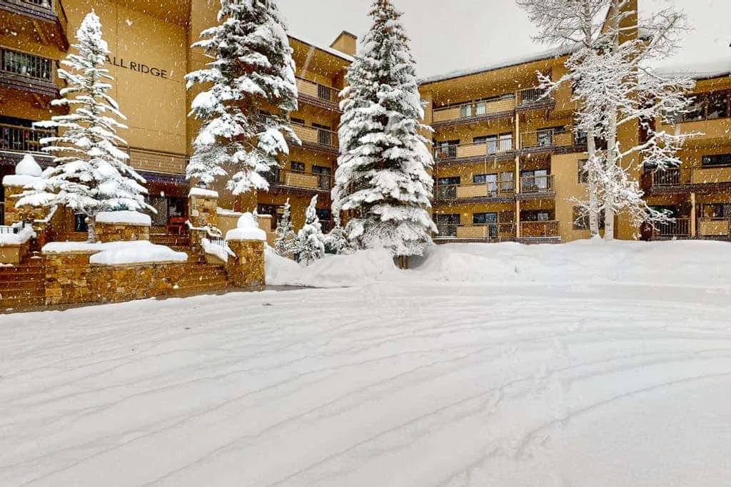 A snow-covered courtyard sits in front of a multi-story building and evergreen trees.