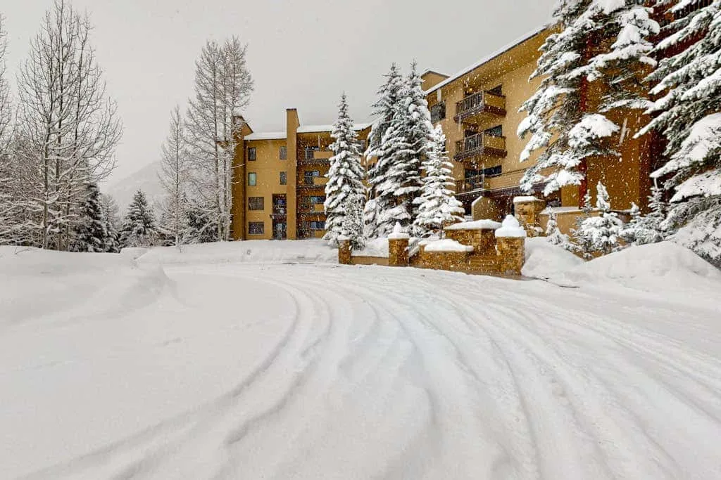 Snow-covered grounds lead toward a multi-story building among evergreen trees.