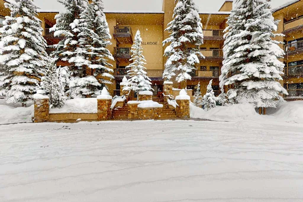 A snow-covered resort building stands behind evergreen trees in a snowy courtyard.