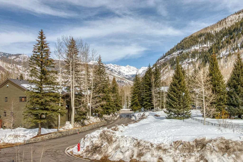 A snowy mountain road curves past trees and houses beneath a partly cloudy sky.