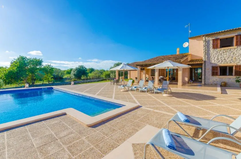 A swimming pool and paved patio sit beside a stone-and-stucco house.