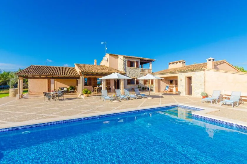 A swimming pool and paved patio sit in front of a low, sunlit building.
