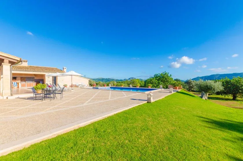 A sunny patio beside a swimming pool overlooks a broad green lawn.