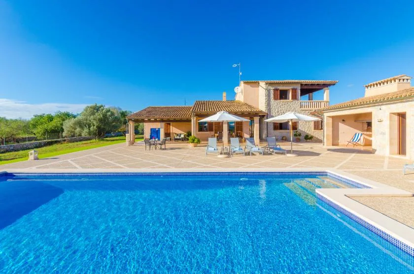 A swimming pool in front of a sunlit villa, with patio seating and umbrellas.