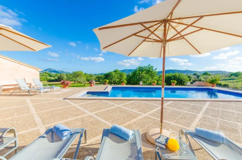 An outdoor pool and sun loungers sit on a paved terrace beneath large umbrellas.