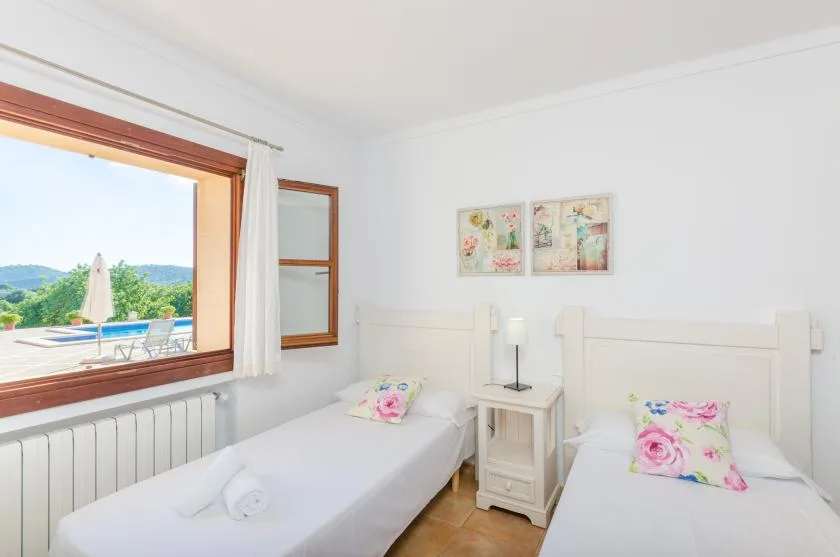 A bright bedroom with two beds and a large open window overlooking greenery.