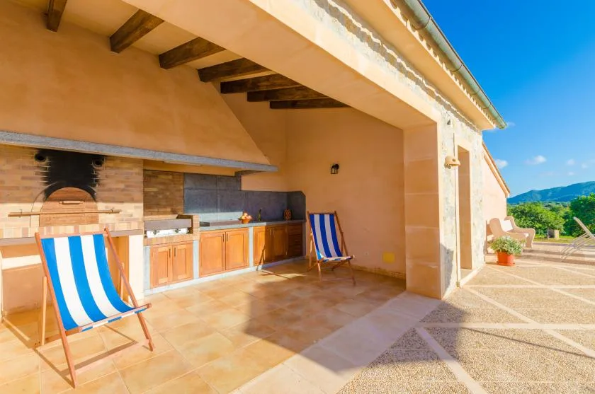 A covered outdoor kitchen opens onto a sunny terrace, with striped seating nearby.