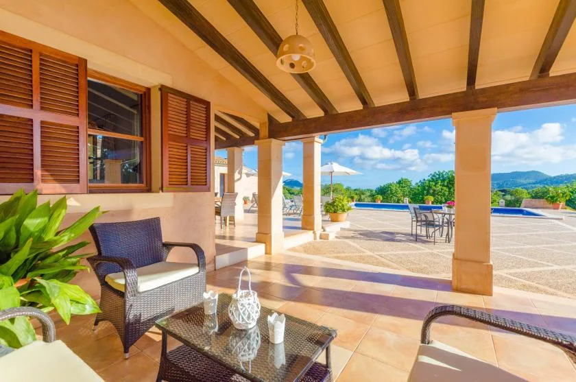 A covered patio opens onto a sunny terrace with a pool and distant hills.