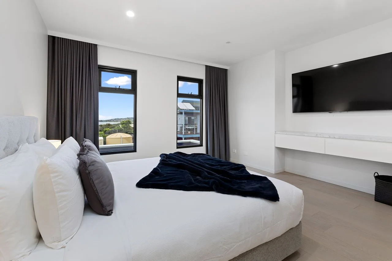 Un dormitorio luminoso con una cama, dos ventanas y un televisor en la pared.