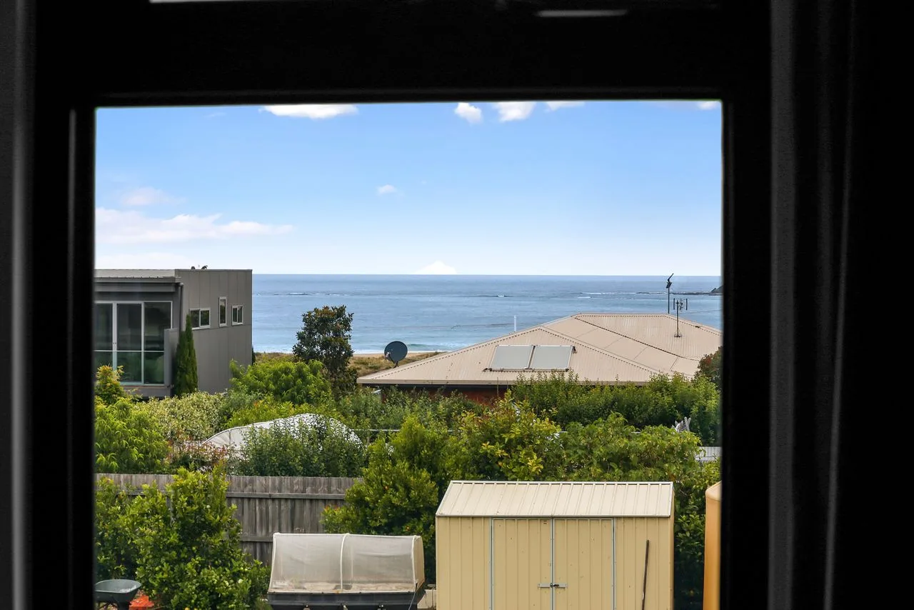 Una ventana da a tejados, vegetación y al océano bajo un cielo despejado.
