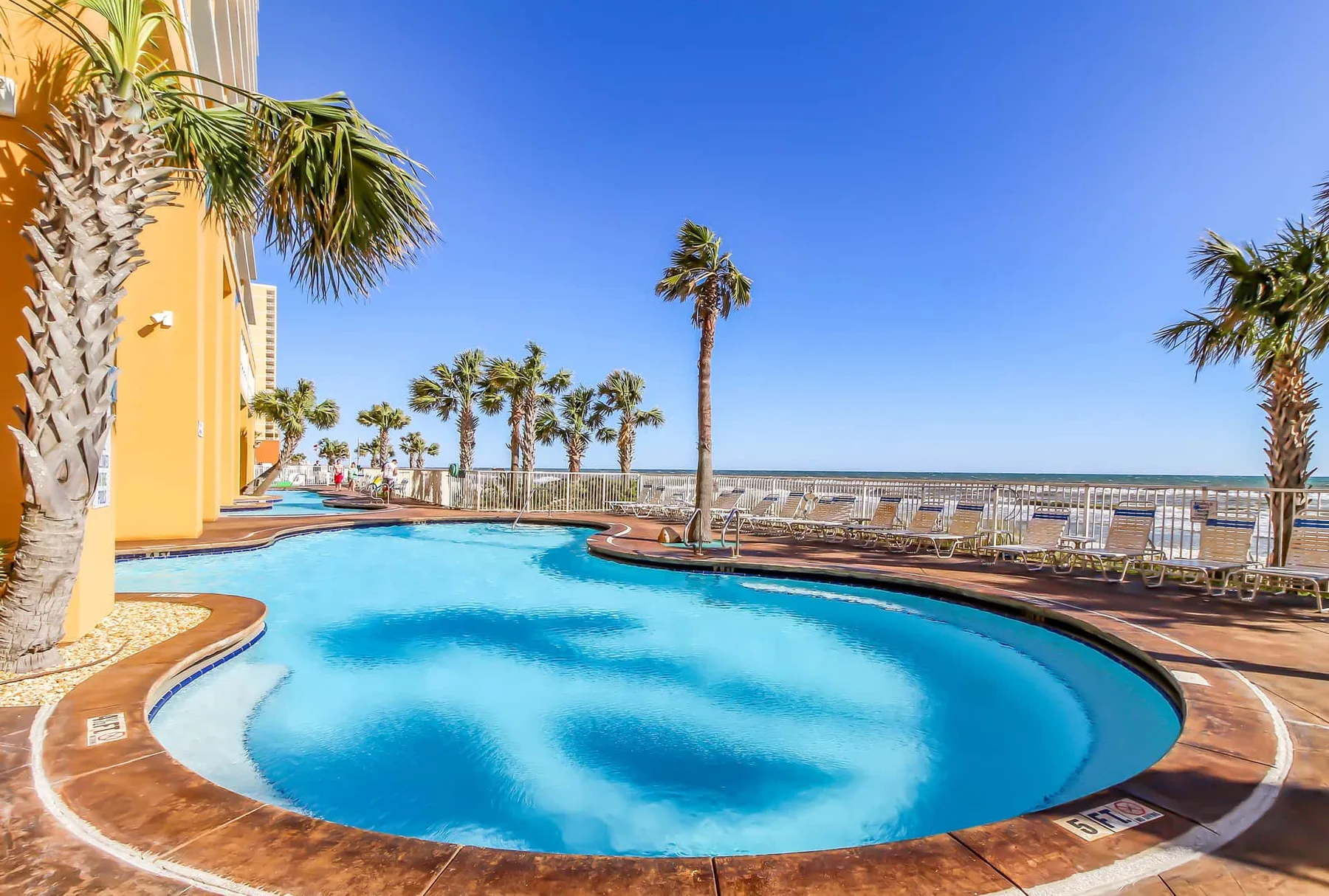 Una piscina exterior curva está junto a un edificio amarillo y palmeras.