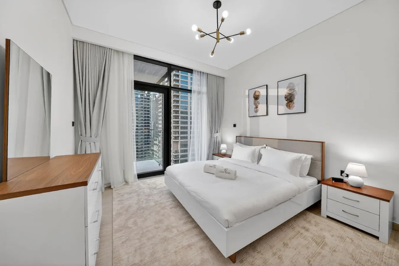 A bright bedroom with a white bed, bedside tables, and large curtained windows.
