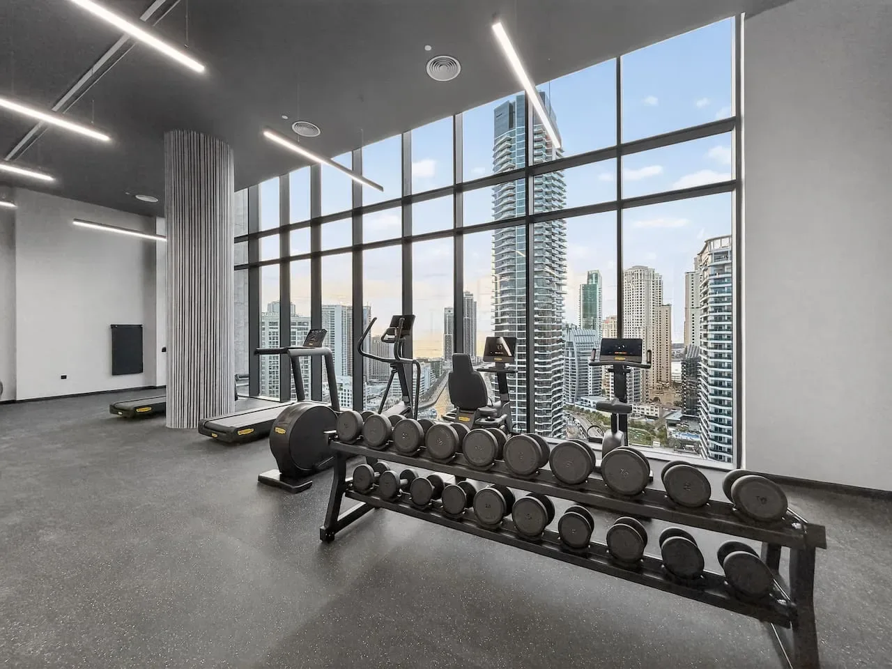 A gym with exercise equipment and weights beside large windows overlooking city buildings.