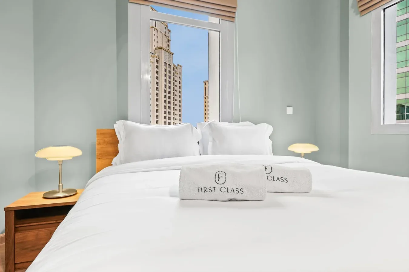 Un dormitorio con una cama tendida, mesitas de noche y ventanas con vista a edificios cercanos.
