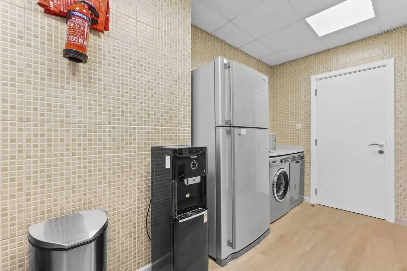 Un cuarto de servicio con azulejos, refrigerador, lavadora y dispensador negro.