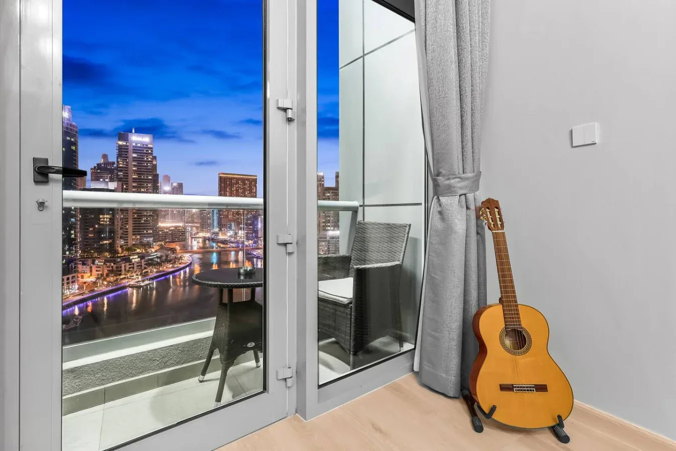 Un balcón con asientos ofrece vistas de edificios iluminados al anochecer, junto a una guitarra en el interior.