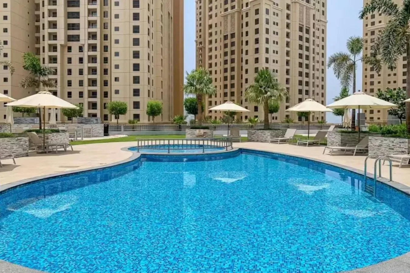 Una piscina al aire libre está rodeada de edificios altos y zonas de descanso con sombra.