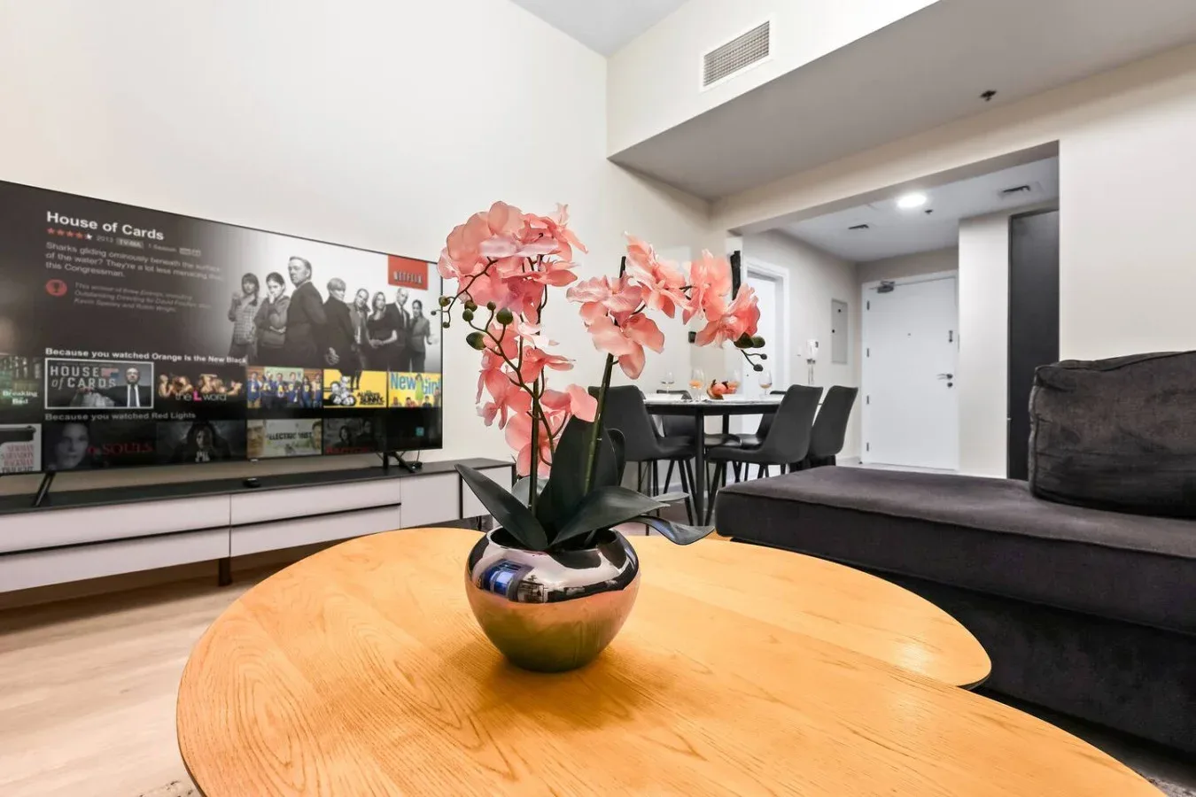 Una sala de estar con televisor, mesa de comedor y flores rosas sobre la mesa de centro.