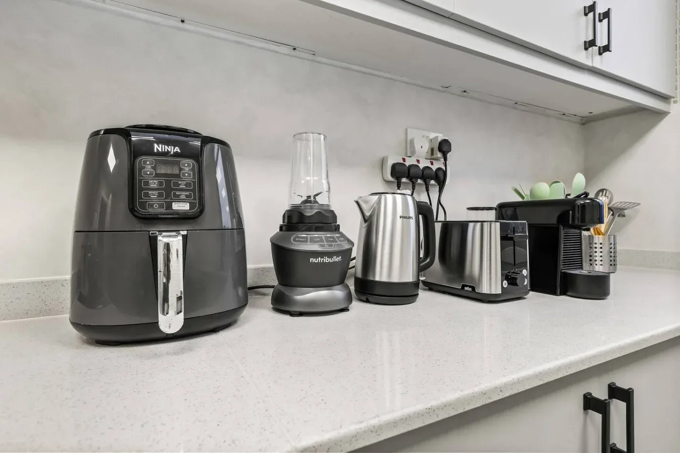 Una encimera de cocina muestra varios electrodomésticos pequeños bajo armarios blancos.