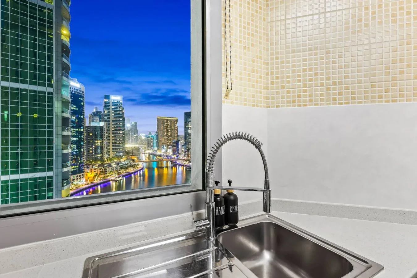 Un fregadero de cocina está junto a una gran ventana con vistas a edificios iluminados al anochecer.