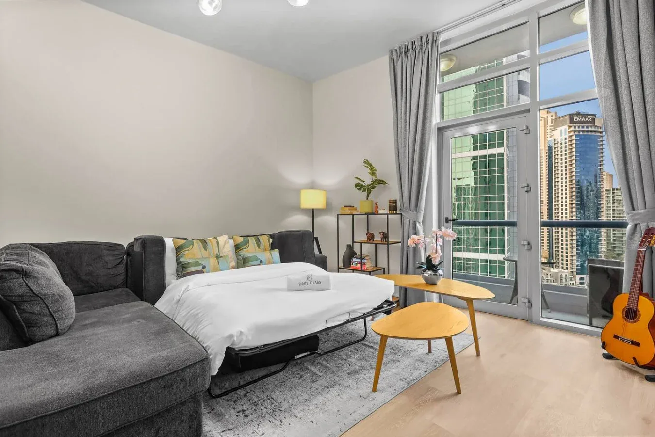 Una sala luminosa con sofá seccional gris, una cama y ventanales.