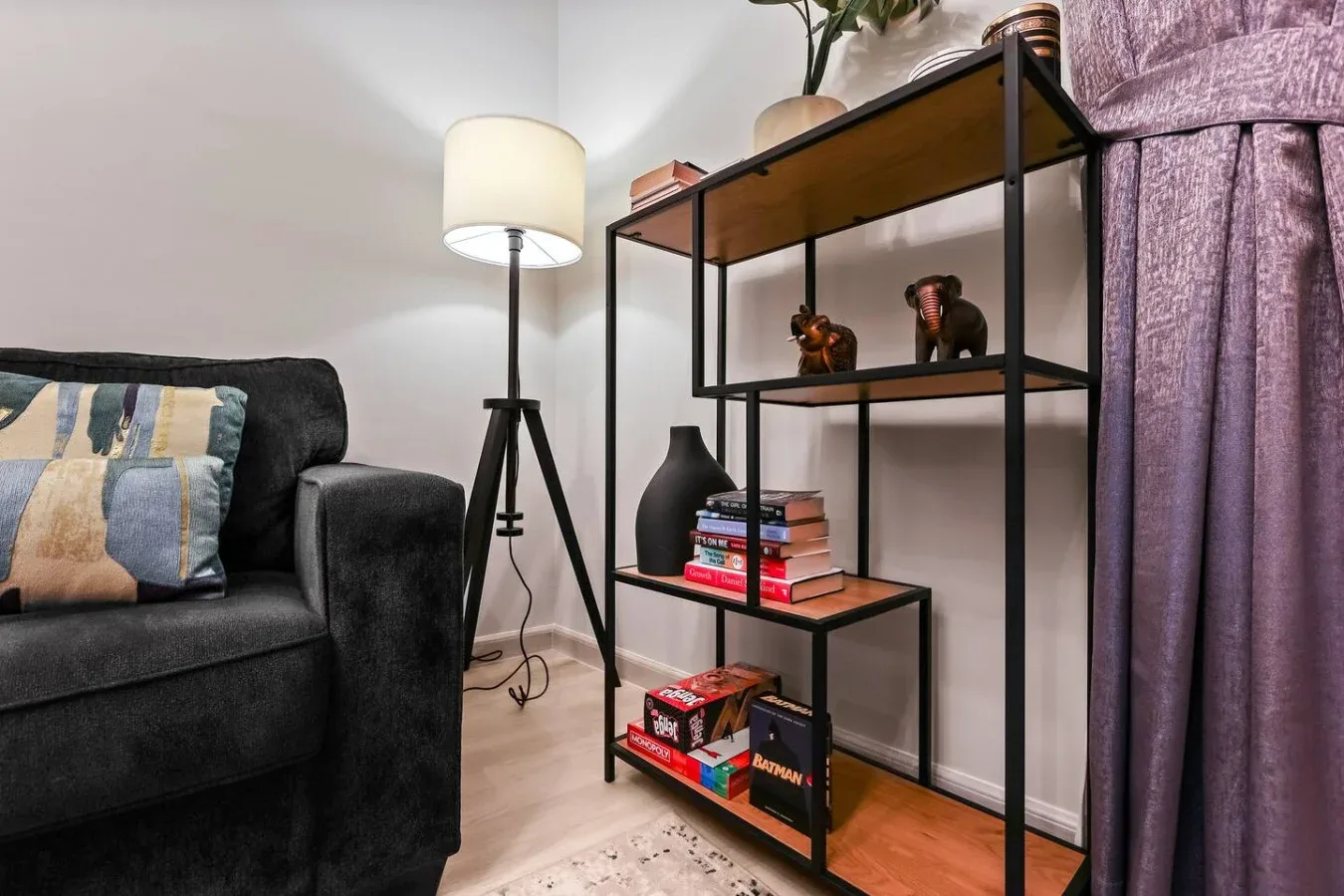 Una sala de estar con un sofá oscuro, una lámpara de pie y estanterías abiertas junto a cortinas moradas.