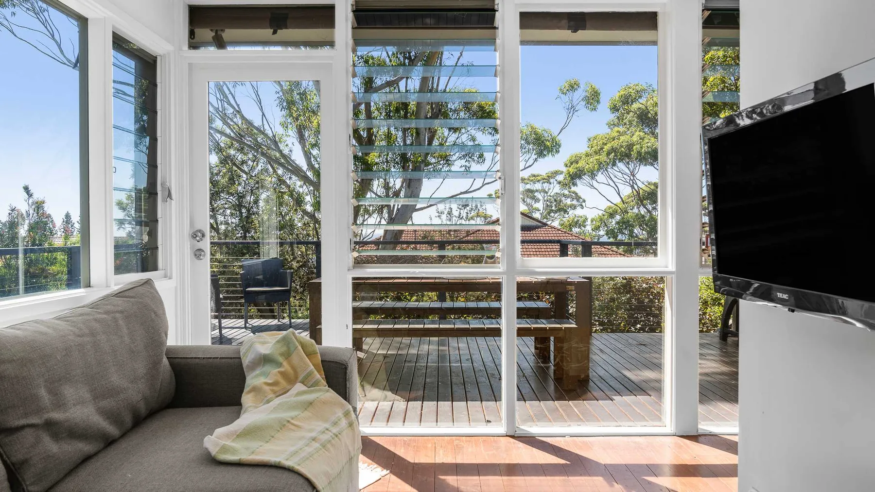Una sala luminosa da a una terraza de madera a través de grandes ventanales.