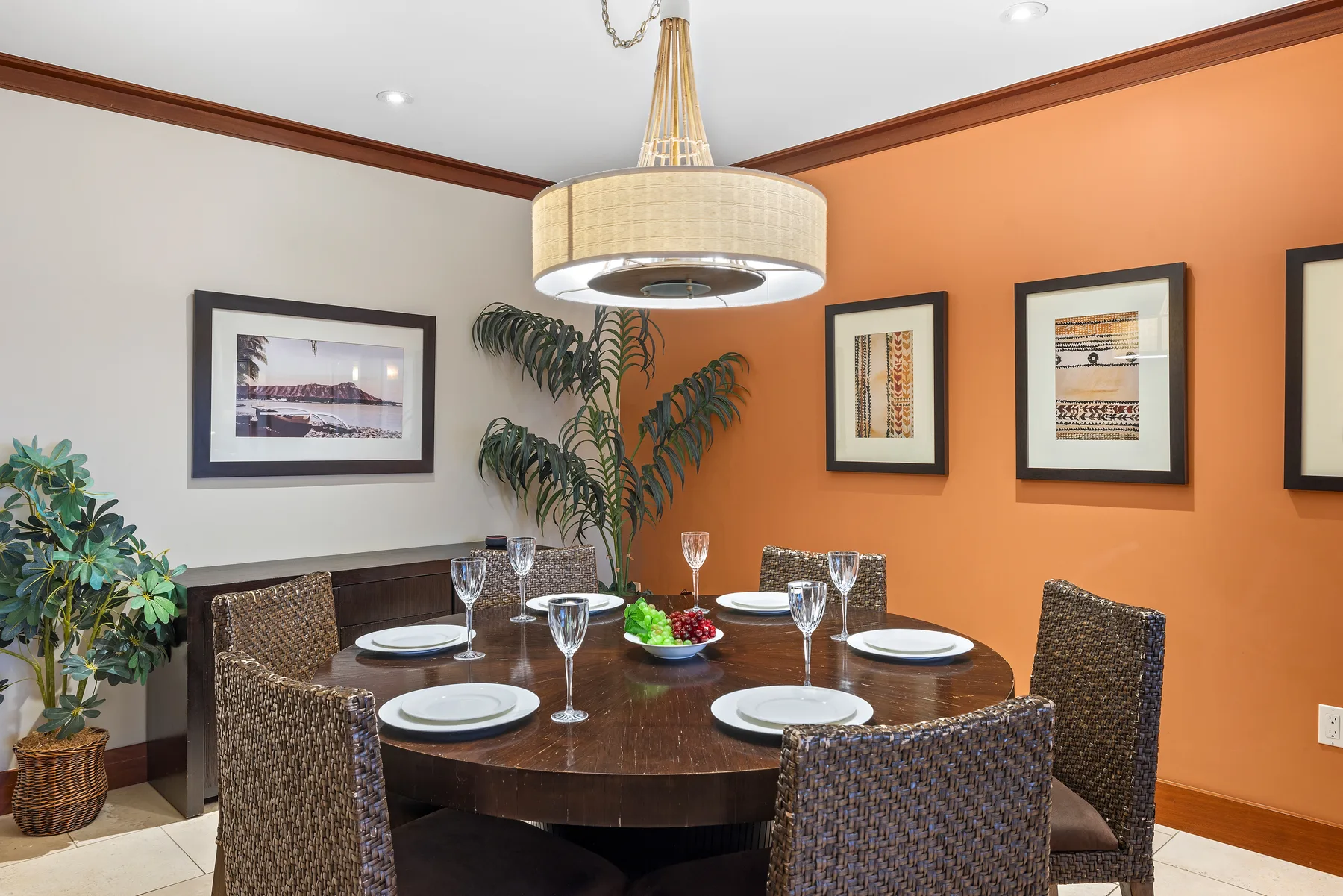 Comedor con una mesa redonda, asientos tejidos y cuadros en una pared naranja.