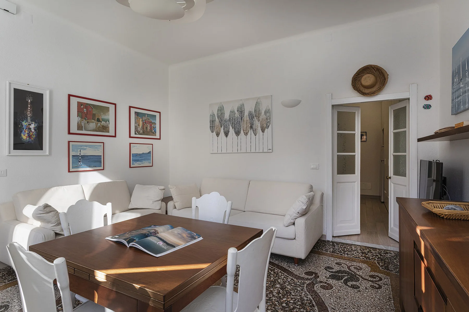 A living and dining room with a wooden table, white seating, and framed wall art.
