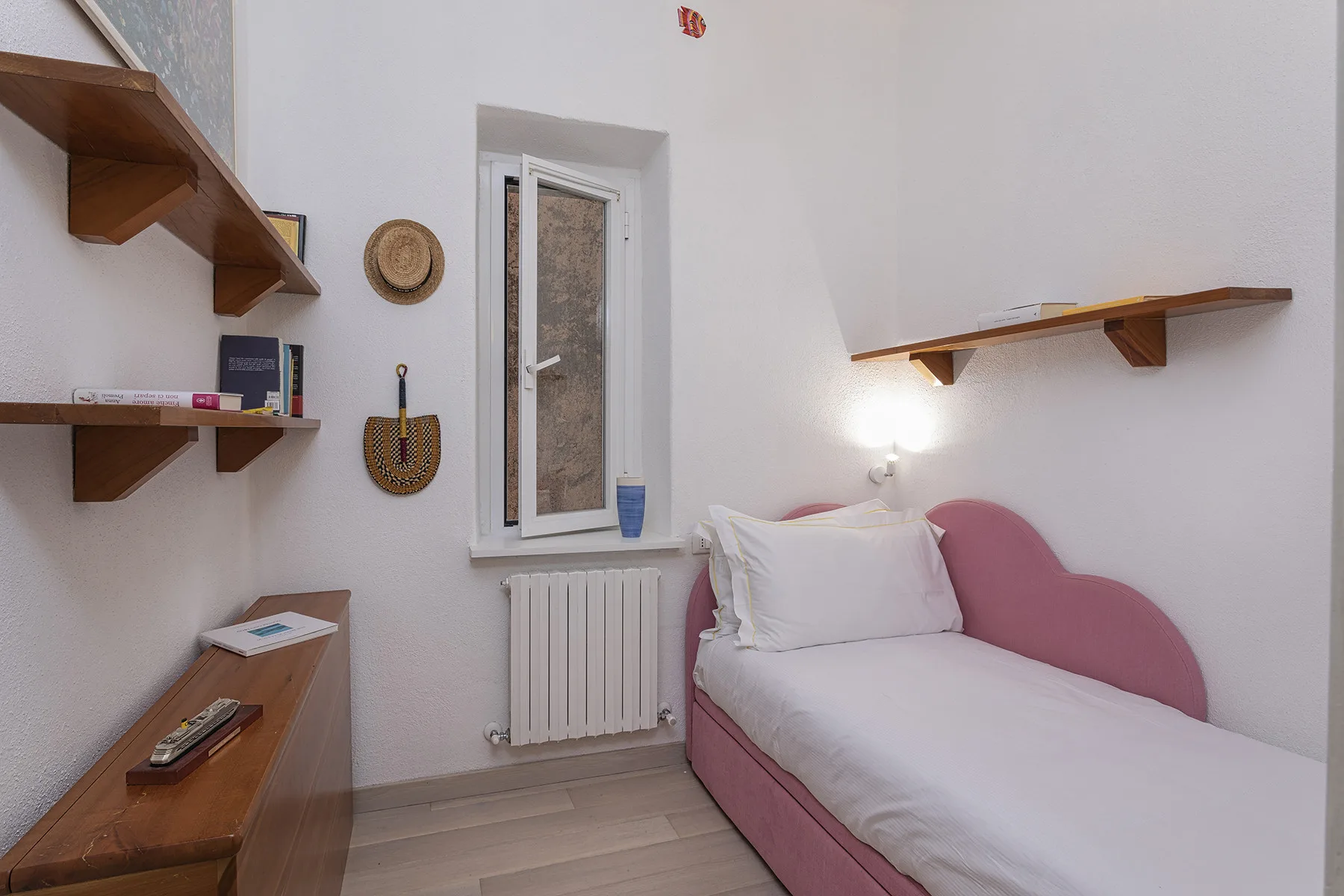 A bedroom with a single bed beneath a wall shelf and beside a window.