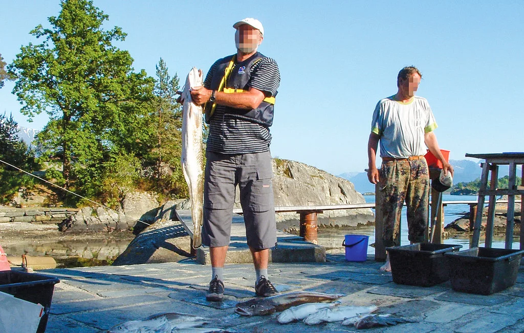 Two people stand on a waterside dock, one holding a large fish.
