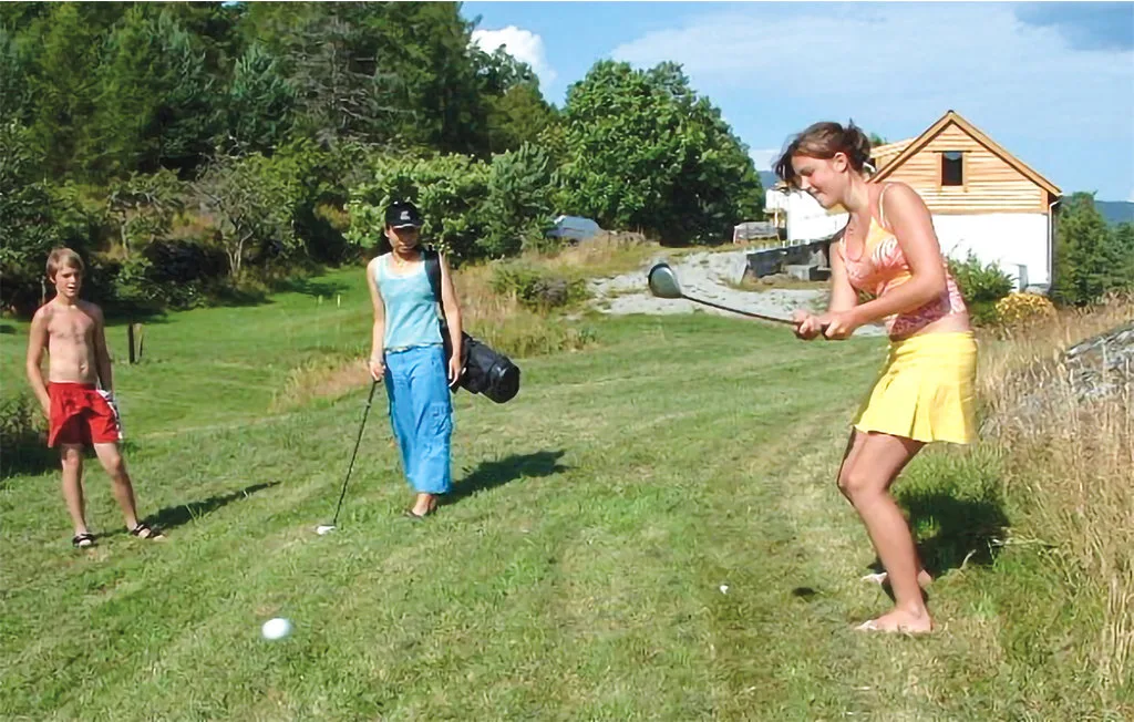 People play golf on a grassy area beside trees and a house.