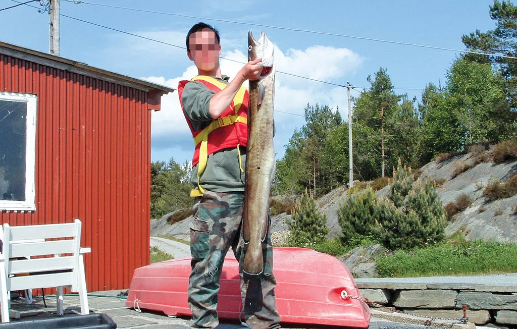 A person holds a large fish beside a red building and an overturned boat.
