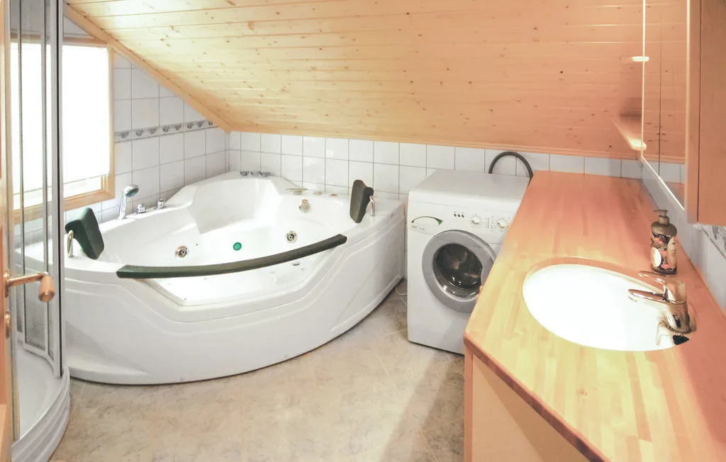 A bathroom with a corner bathtub, washing machine, and sink beneath a sloped wooden ceiling.